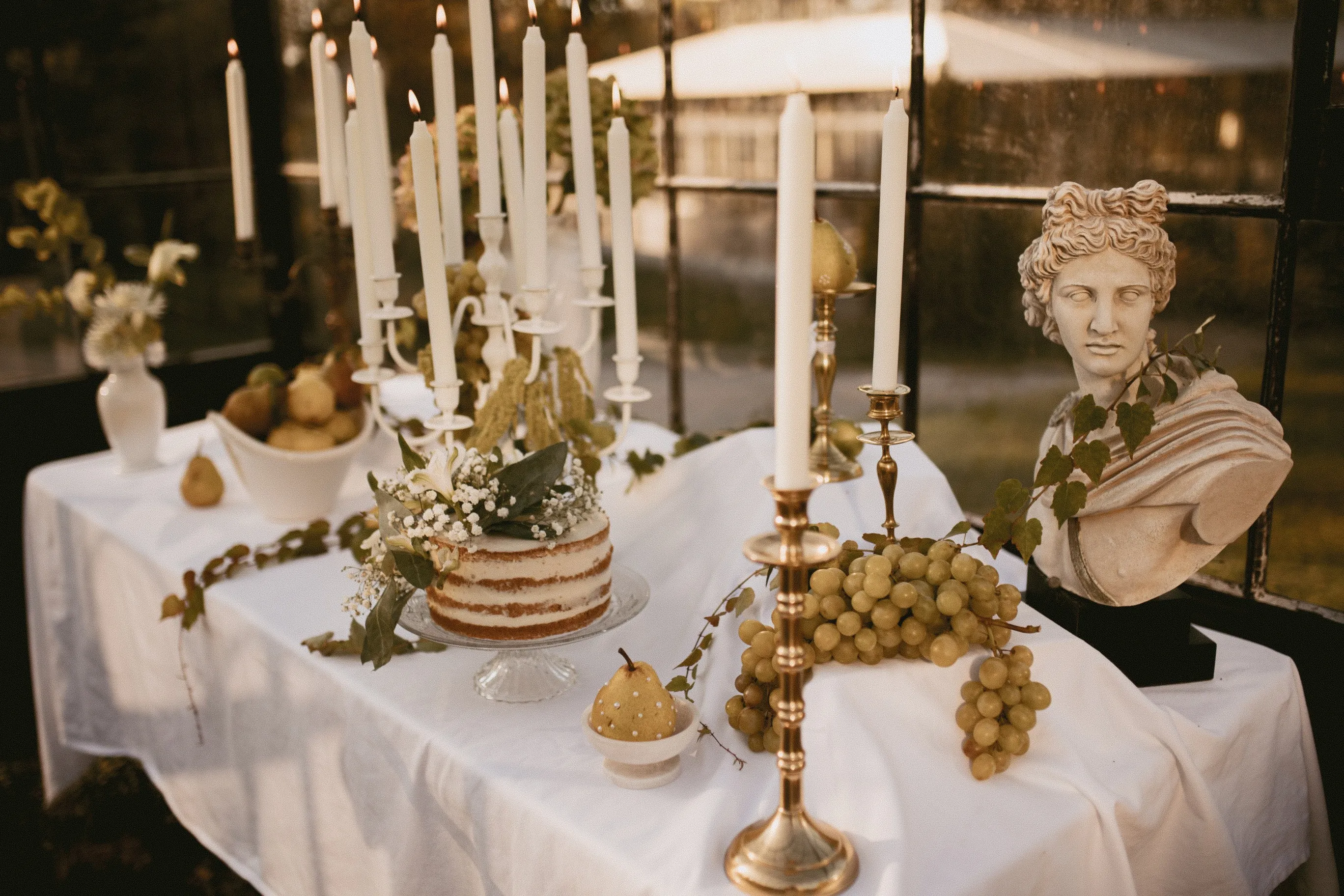 Table de mariage dressée sous une verrière : bougies blanches, gâteau nu, raisins, poires et buste à l'antique